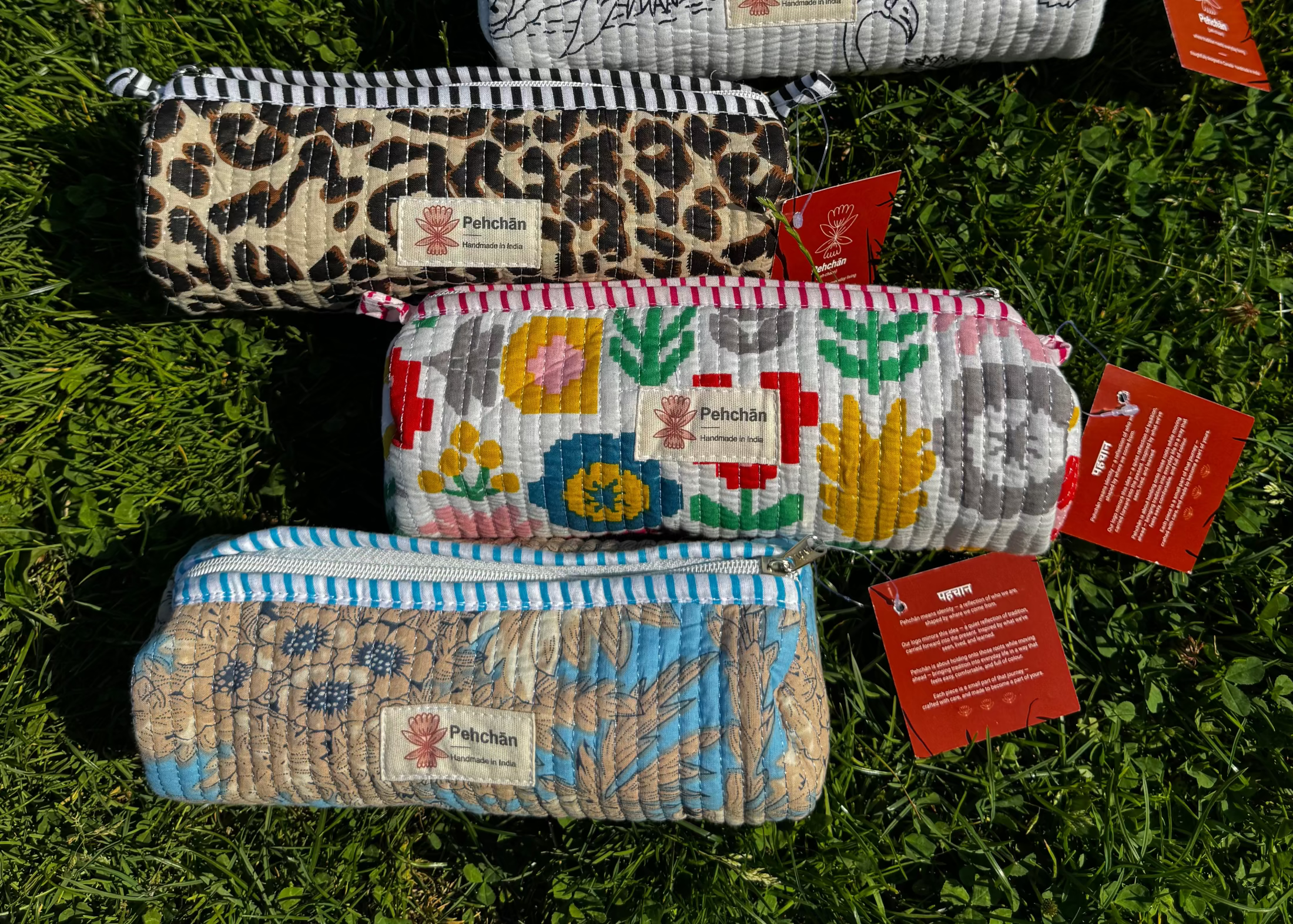 Five quilted fabric pencil cases with various patterns (gingham, floral, leopard, multicolor flowers, and blue tropical print) are arranged on a green grass background. Each case has a white label with the brand name 'The Patch' and red hang tags.