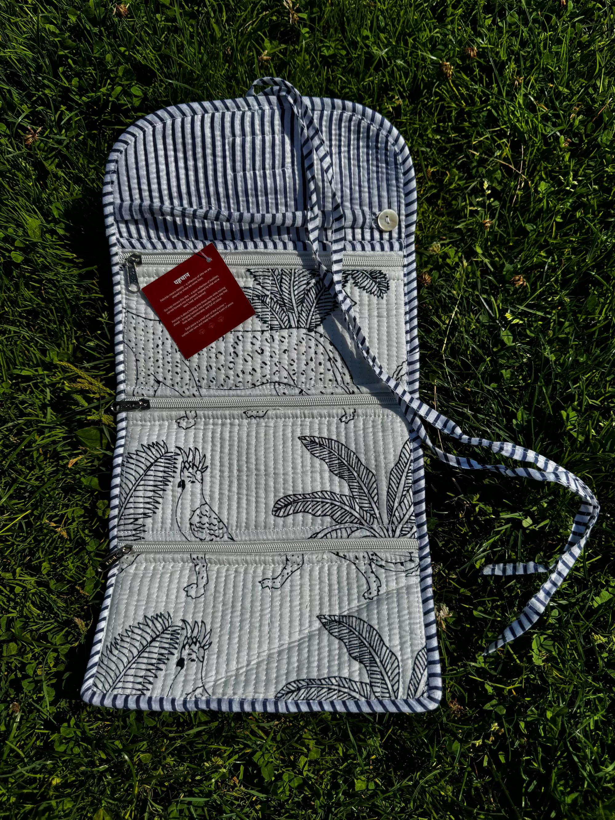 A quilted outdoor blanket with a blue and white striped border, black botanical leaf patterns, and a striped carrying strap, laid flat on green grass. A small red tag is attached to the corner.