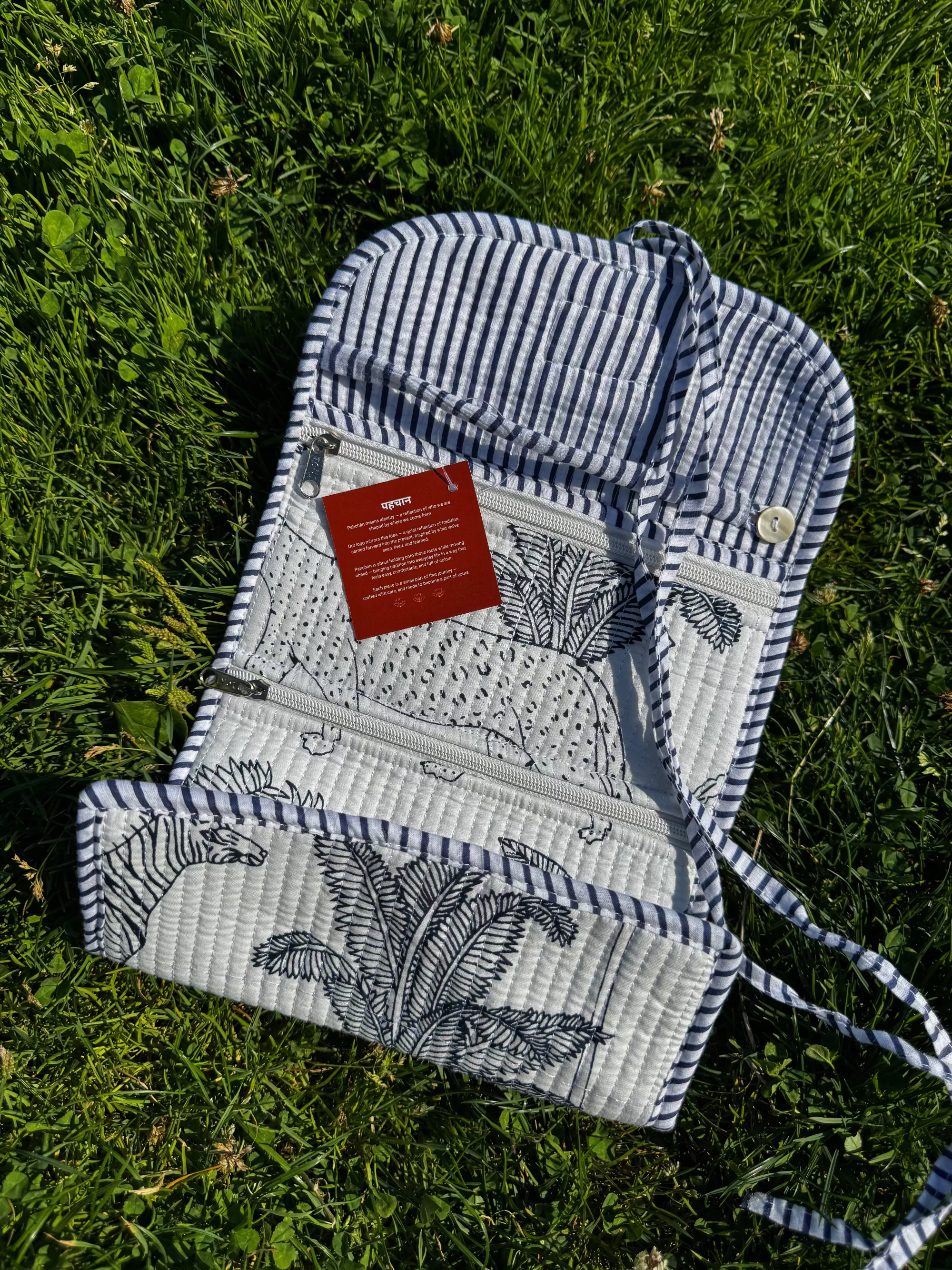 A quilted zippered pouch with a blue and white striped border and black botanical line art, laid flat on green grass. A red paper tag is attached to the zipper.