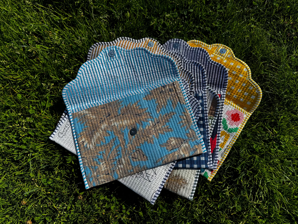 A stack of handmade fabric pouches or small bags with scalloped edges, laid on green grass. The fabrics show different patterns including blue-and-white stripes, a distressed blue-and-brown print, navy-and-white gingham, and a yellow-and-white gingham with a red flower motif.
