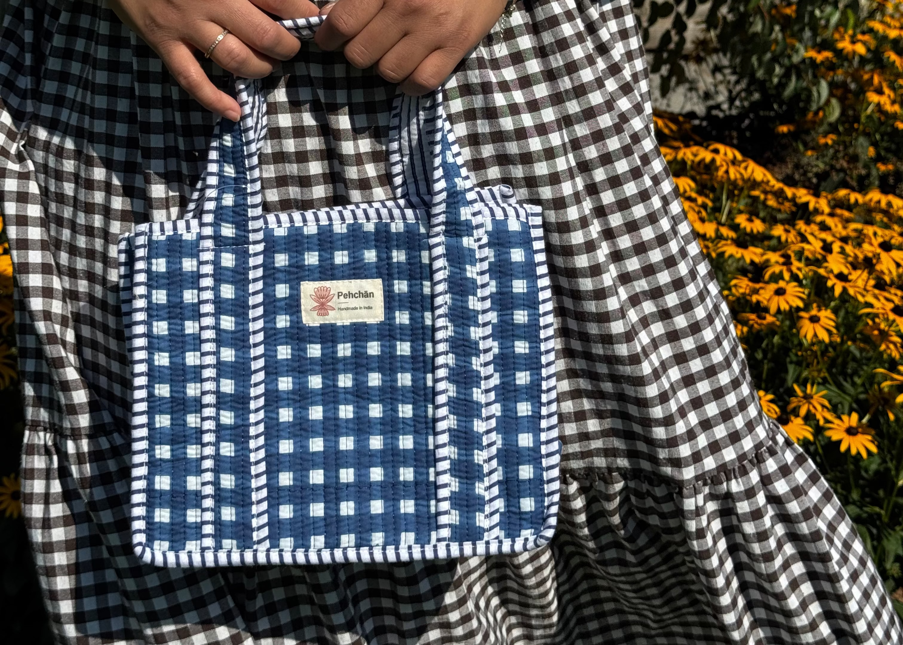 A person wearing a black and white gingham dress holds a blue and white gingham fabric tote bag with white braided handles and trim, set against a background of yellow flowers and green foliage.