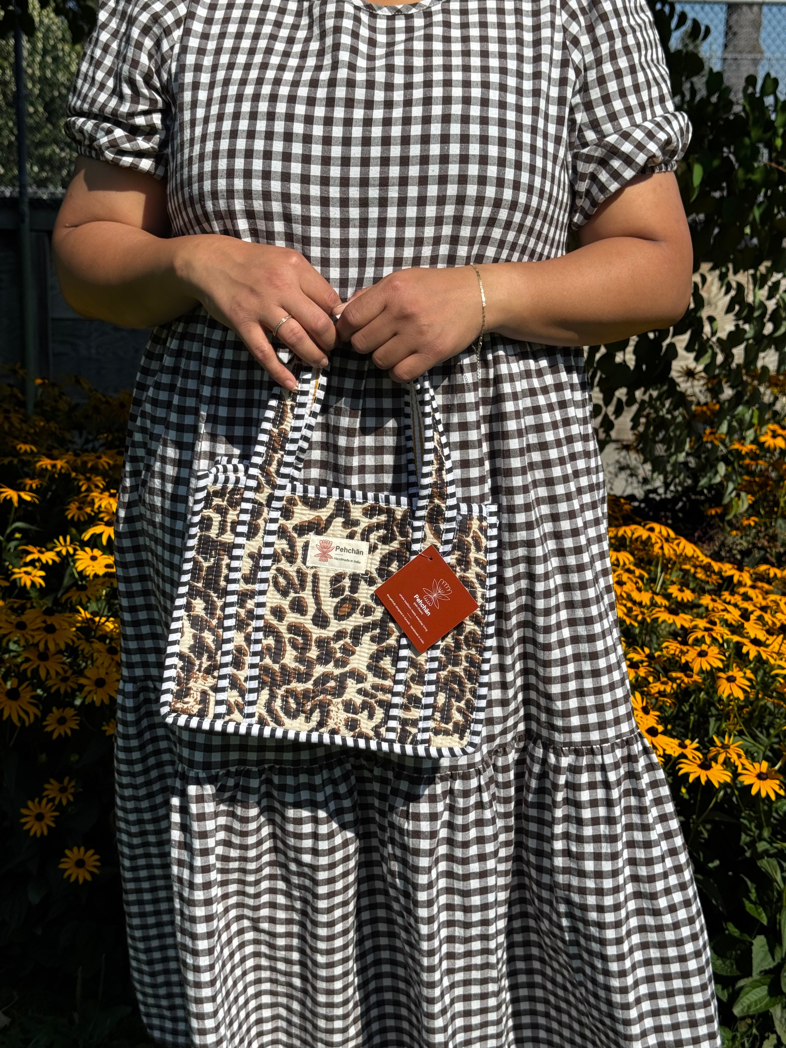 A person wearing a black and white gingham dress holds a small tote bag with a leopard print pattern and black-and-white striped trim, set against a background of yellow flowers and green foliage.