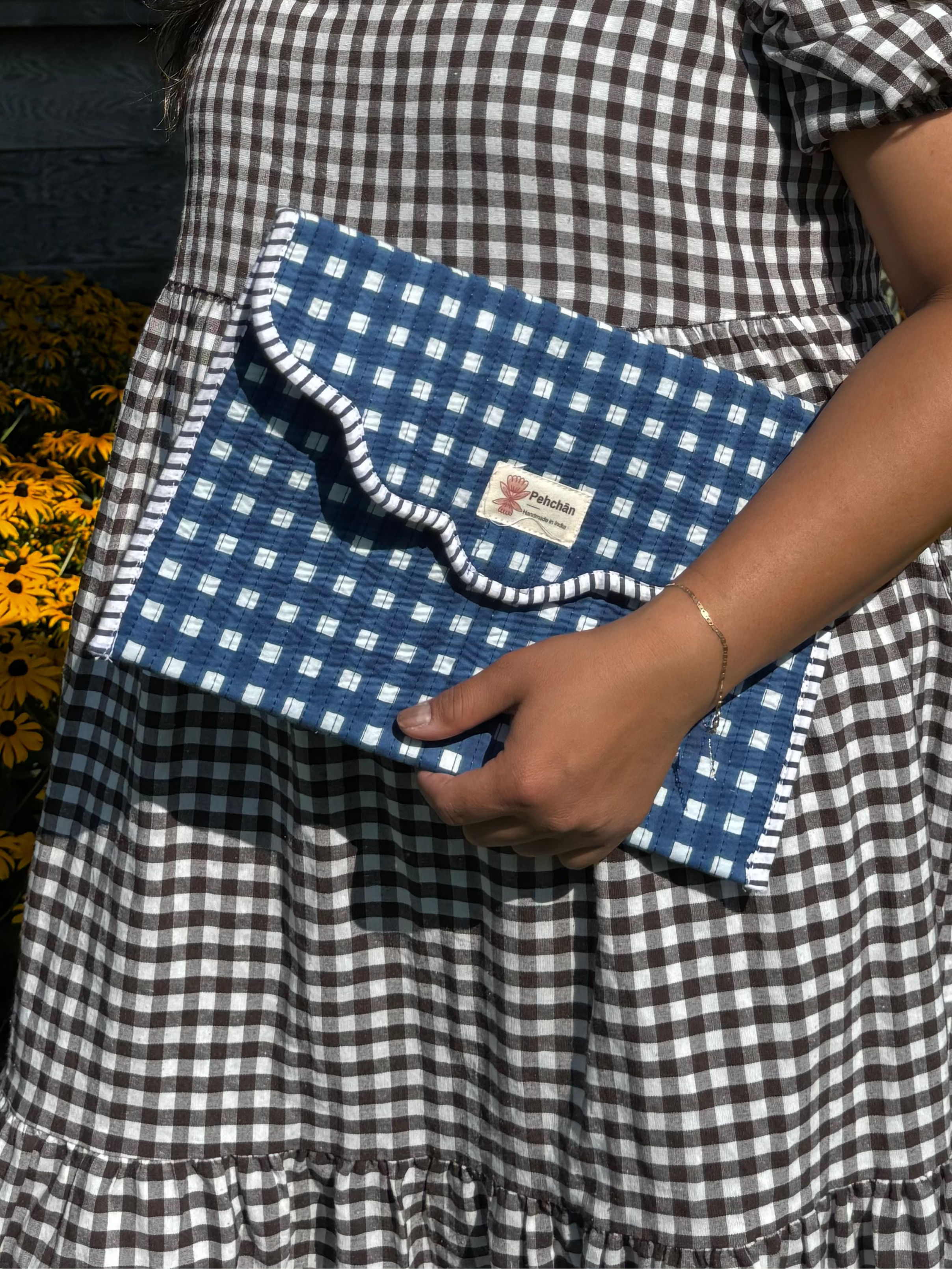 A woman wearing a black and white gingham dress stands outdoors holding a matching blue and white gingham clutch bag with a ruffled trim.