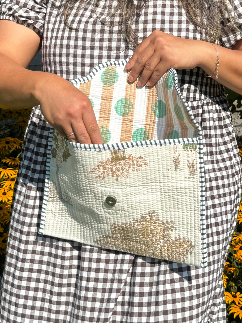 A woman wearing a black and white gingham dress stands outdoors in front of yellow flowers and a tree, holding and opening a small quilted clutch bag with a striped lining.