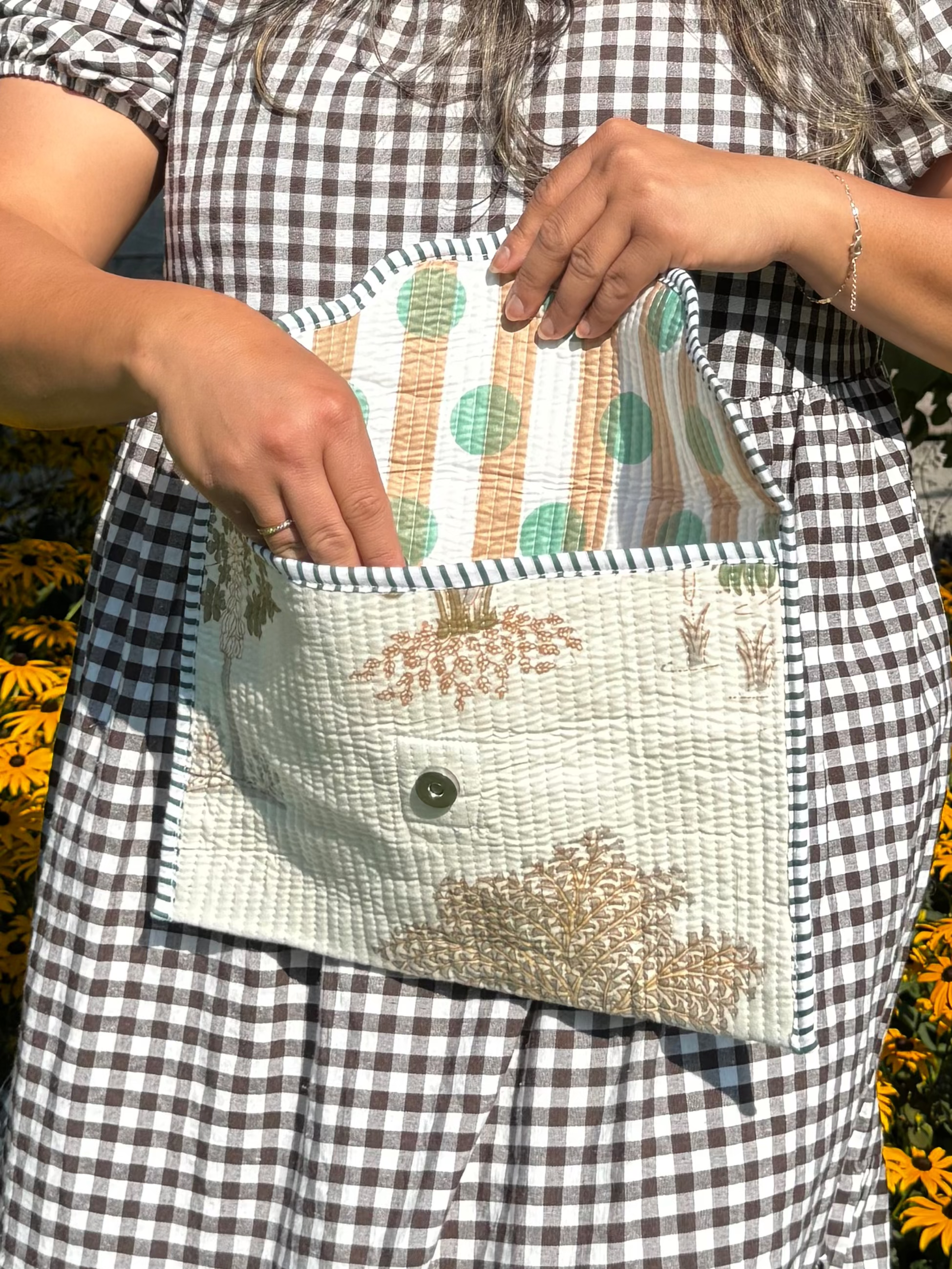 A woman wearing a black and white gingham dress stands outdoors in front of yellow flowers and a tree, holding and opening a small quilted clutch bag with a striped lining.