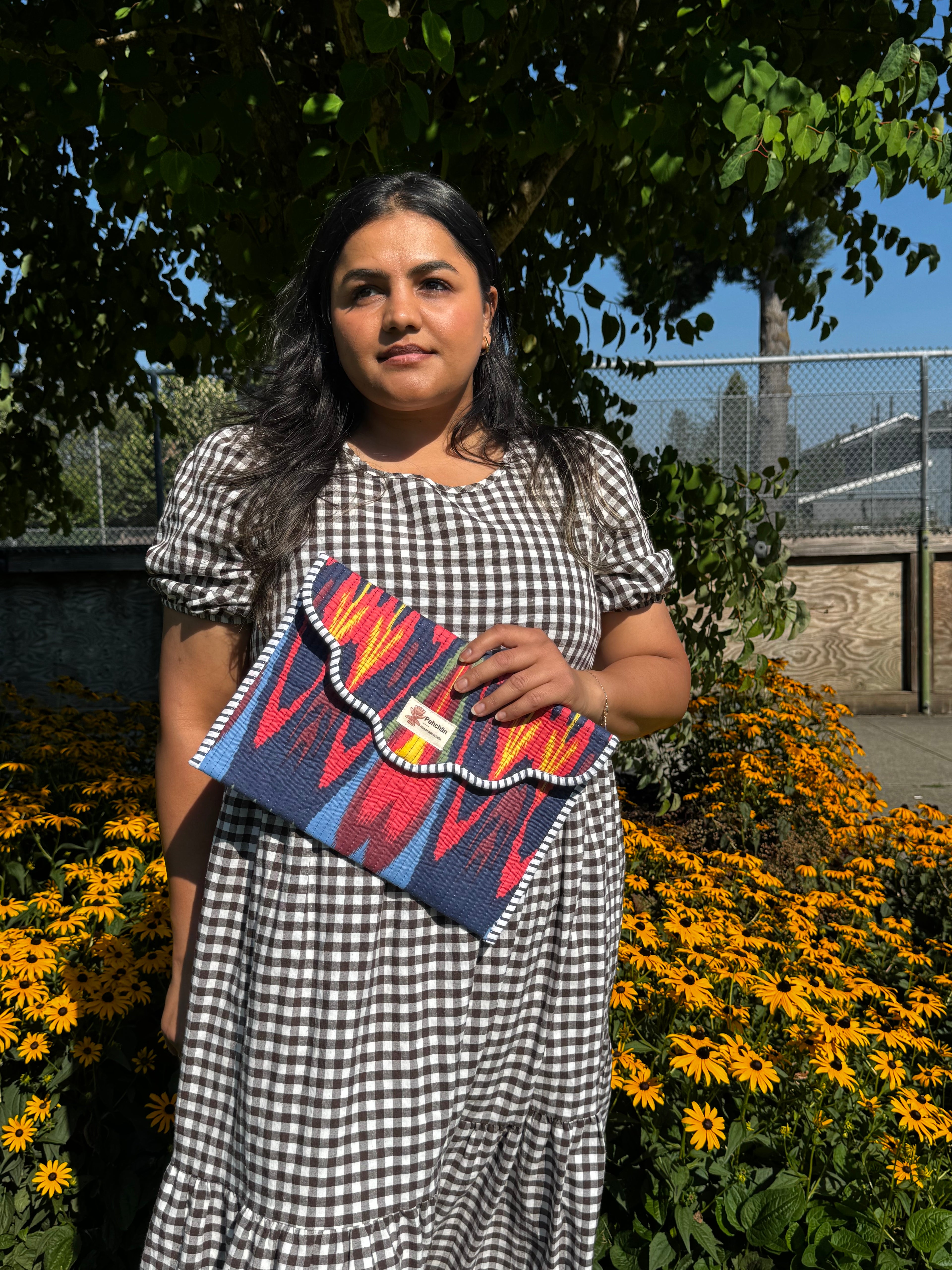 A woman wearing a black and white gingham dress stands outdoors in front of yellow flowers and green foliage, holding a colorful patterned clutch bag.