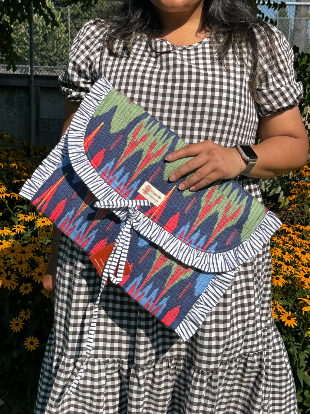 A woman wearing a black and white gingham dress stands outdoors in a garden holding a colorful patterned clutch bag with a white fringed border.