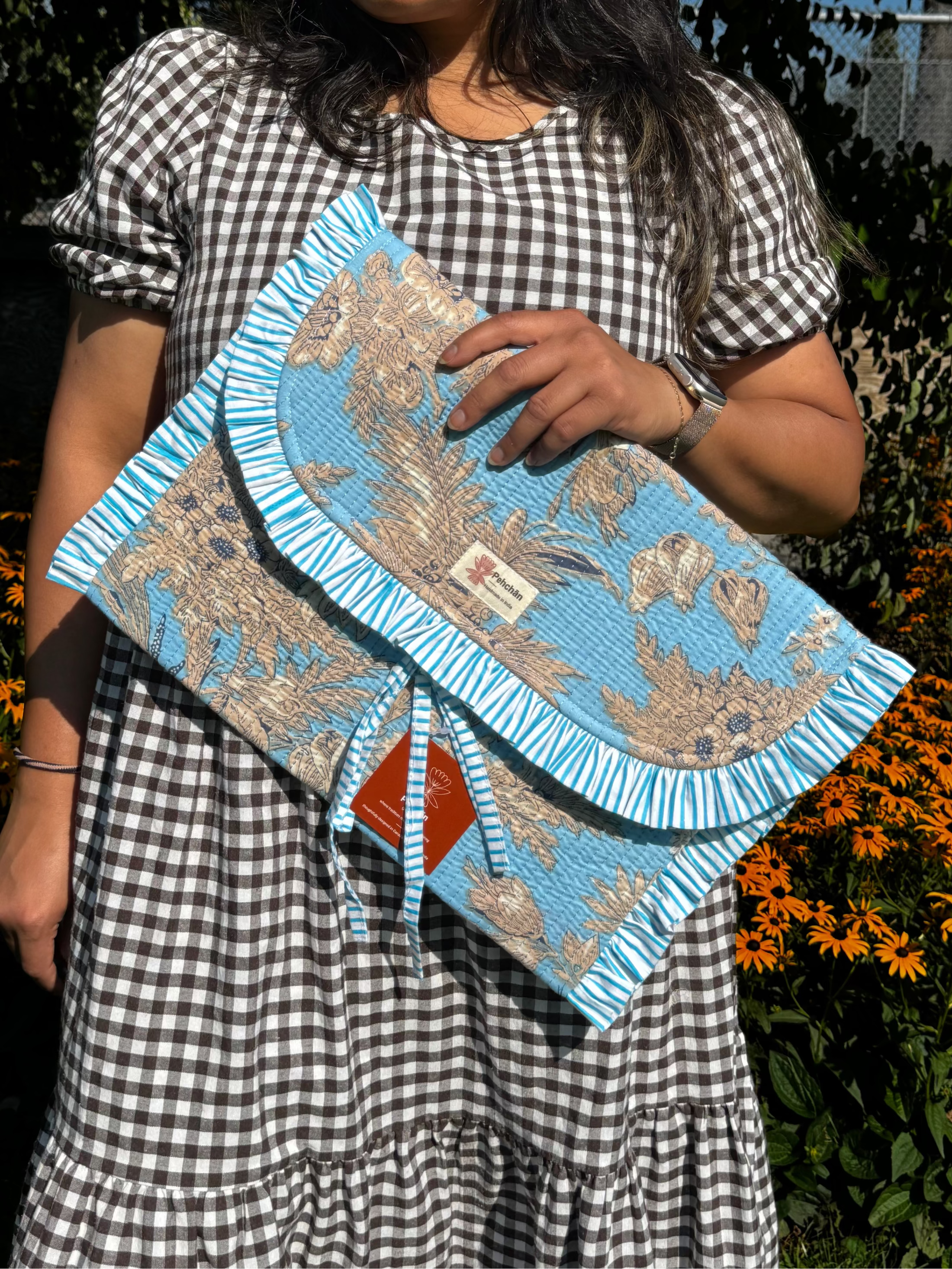 A woman wearing a black and white gingham dress holds a light blue and beige patterned clutch bag with a fringed edge in an outdoor garden setting.