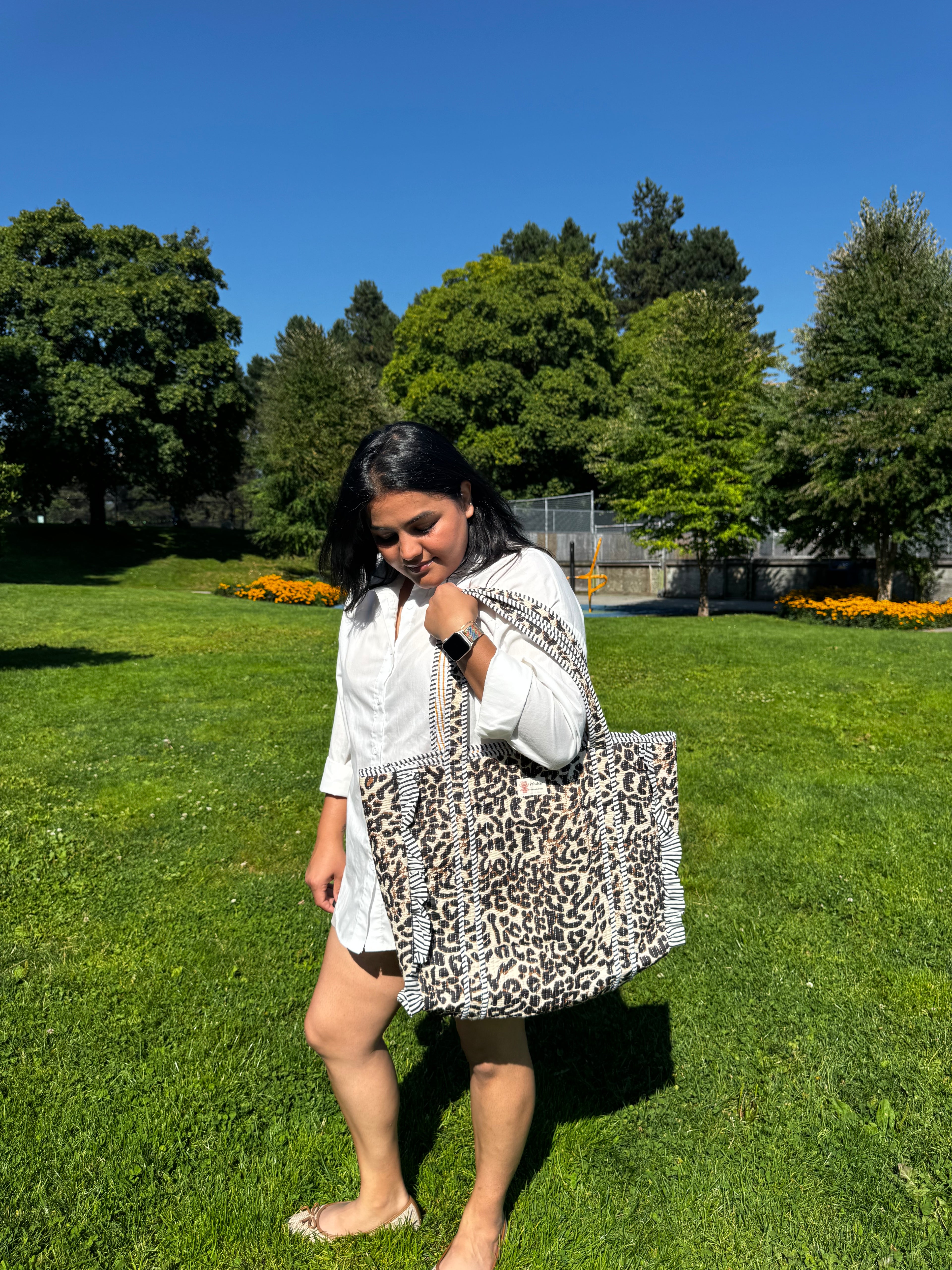 A woman in a white shirt and shorts walks on a green lawn carrying a large leopard-print tote bag under a clear blue sky with trees in the background.