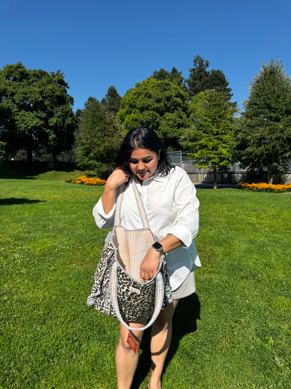 A woman wearing a white button-up shirt and gray shorts stands on a green grass lawn under a blue sky, holding a leopard-print shoulder bag with a striped top handle and white beaded fringe.