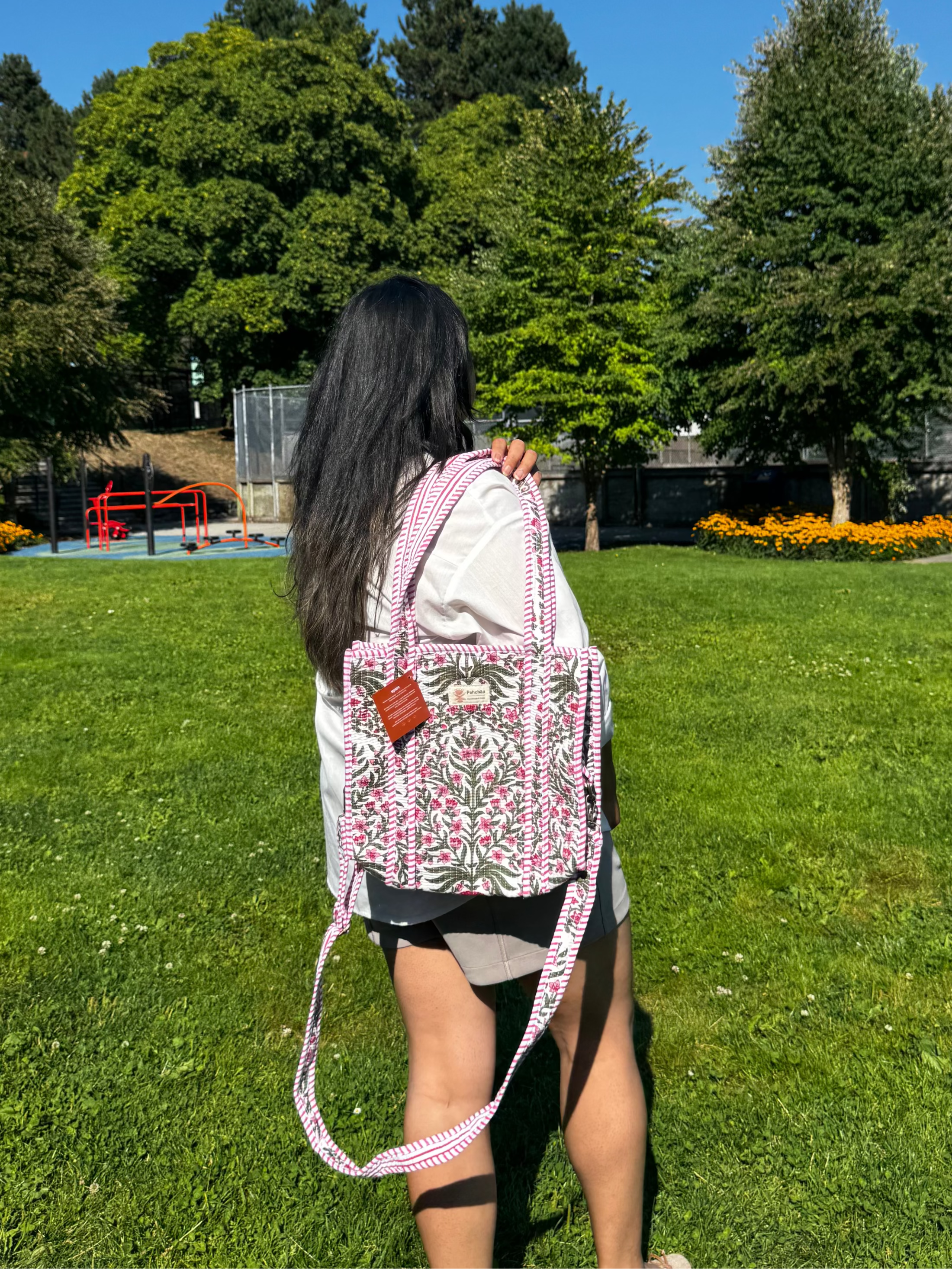 A woman seen from behind wearing a light-colored jacket and shorts, carrying a patterned pink and white tote bag with a brown leather patch on a green grassy park lawn under a blue sky.