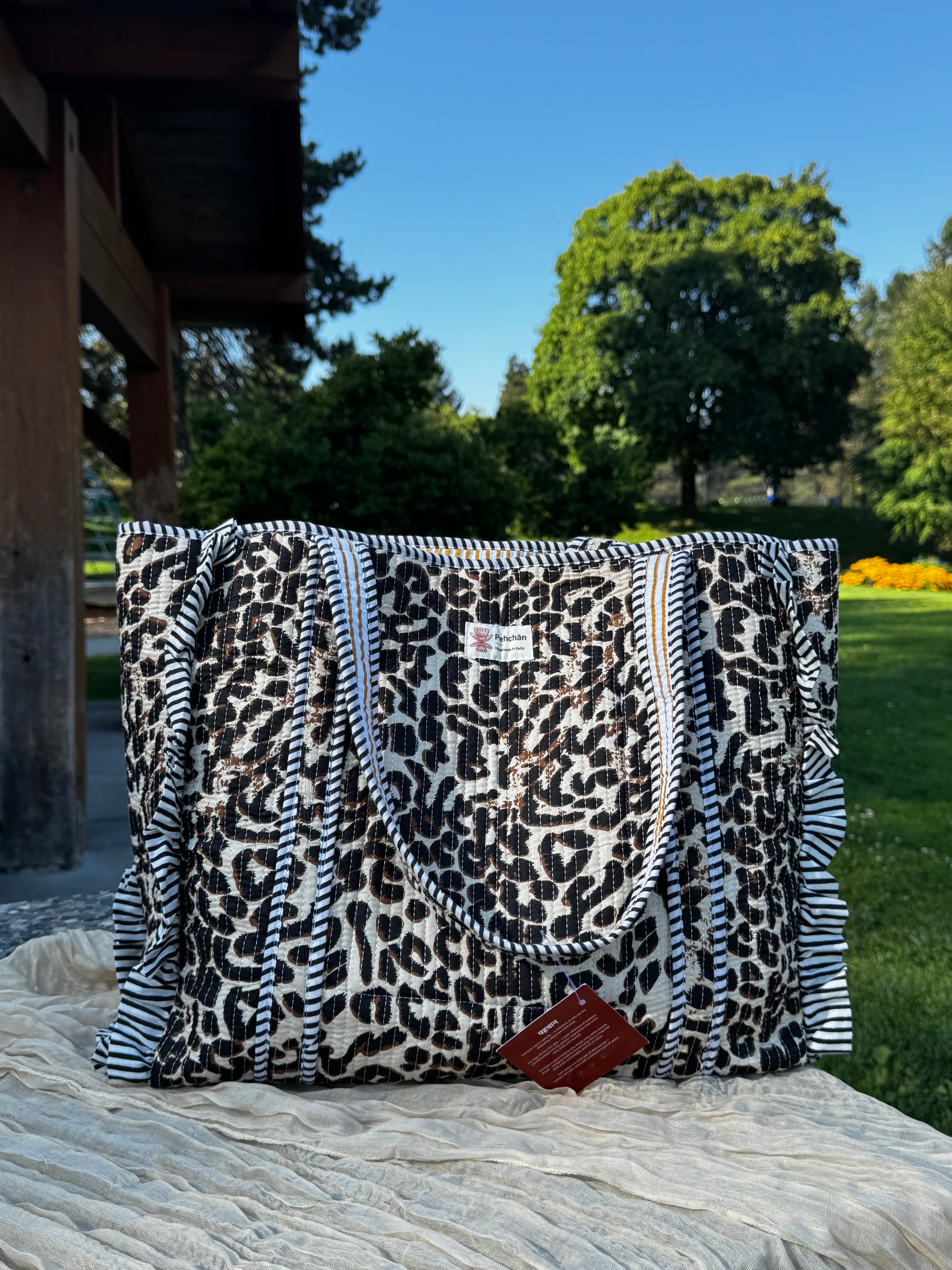 A large tote bag with a black and white leopard print pattern and striped handles, sitting outdoors on a textured blanket with a garden and wooden structure in the background.