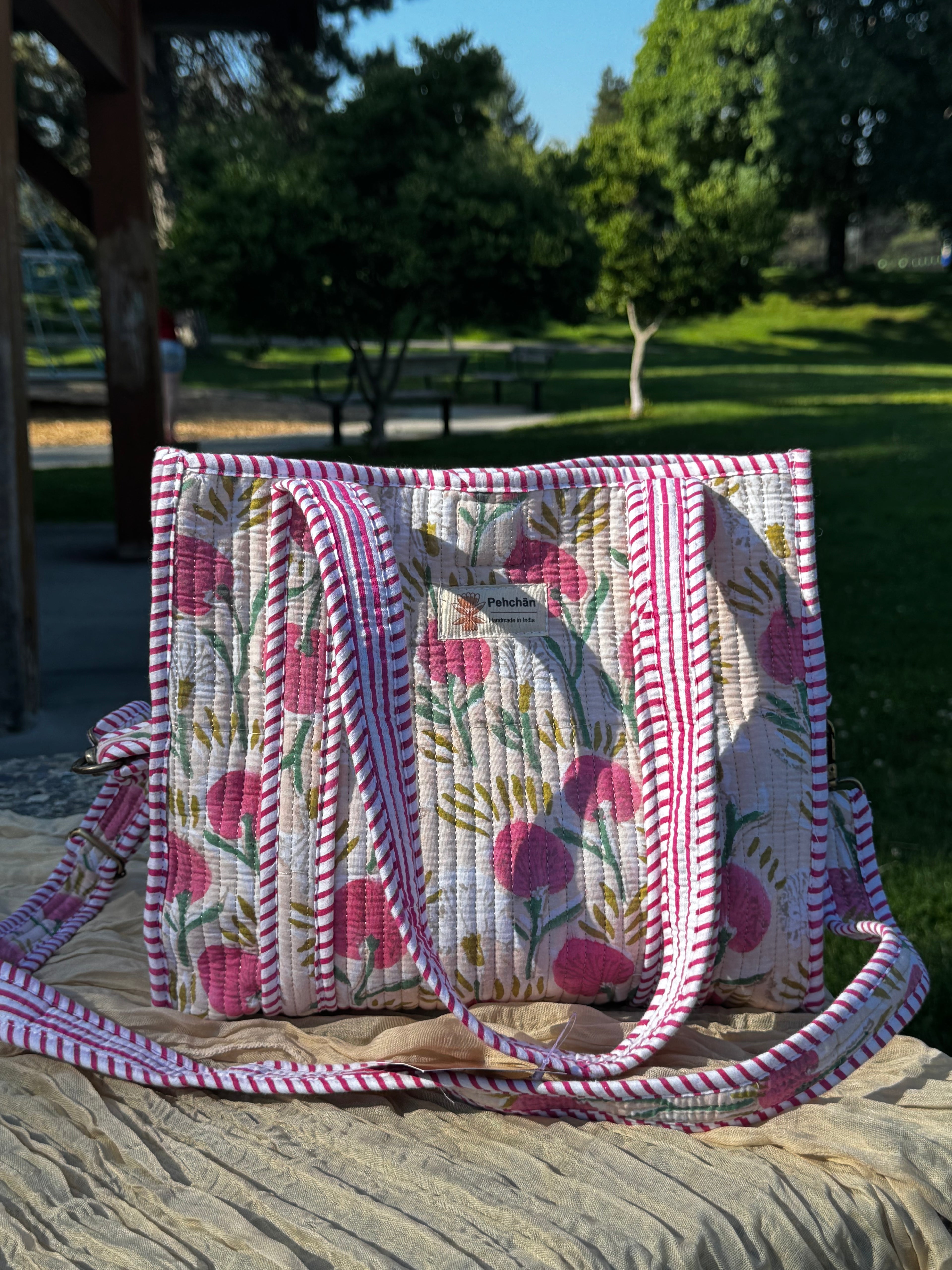 A quilted shoulder bag with a pink and white floral pattern and striped borders, resting on a textured blanket in an outdoor park setting.