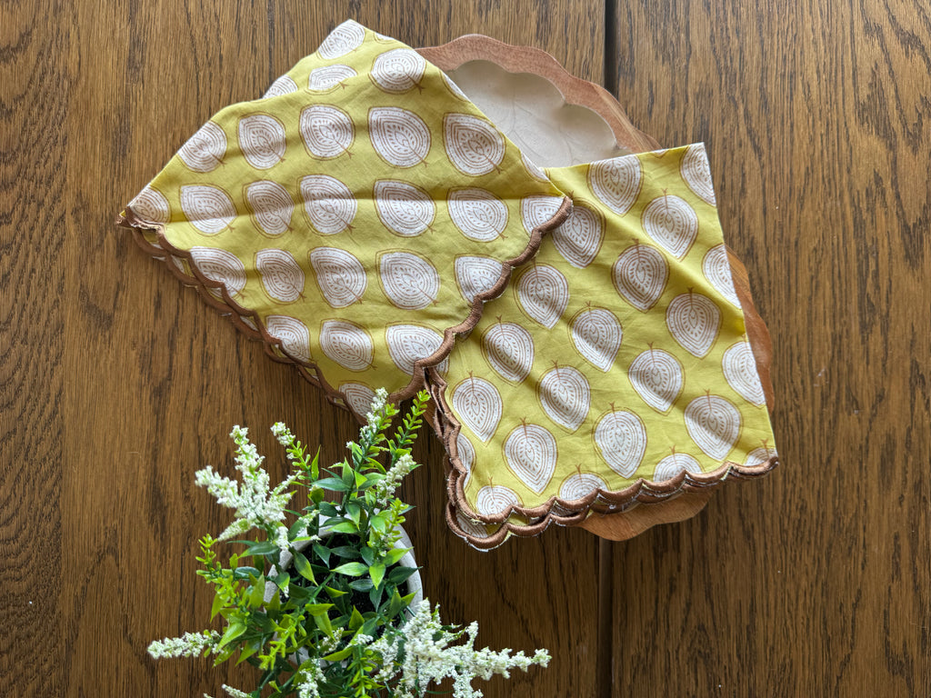 Two square yellow napkins with white printed paisley motifs and scalloped edges, arranged on a wooden surface next to a small green plant.