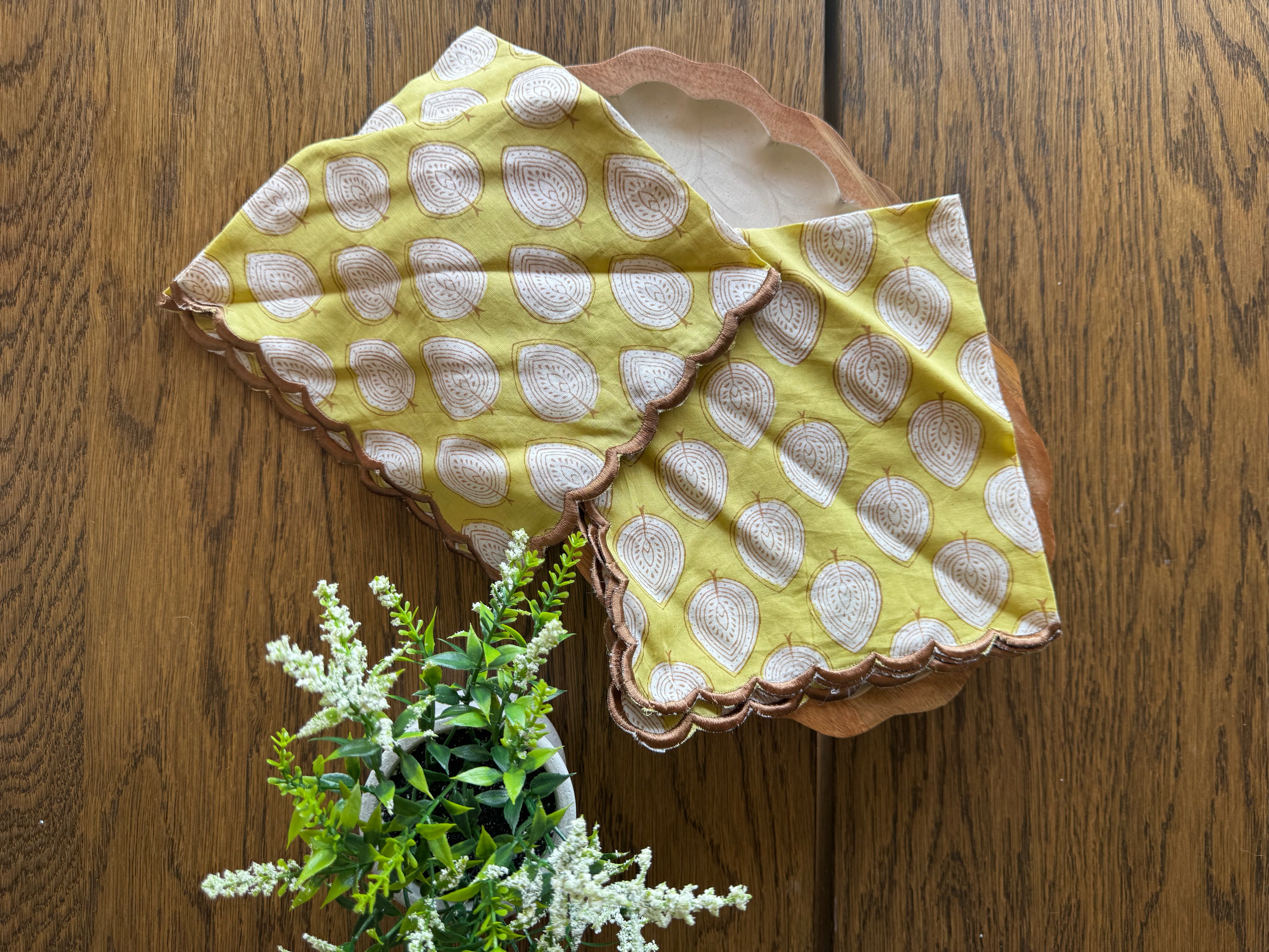 Two square yellow napkins with white printed paisley motifs and scalloped edges, arranged on a wooden surface next to a small green plant.