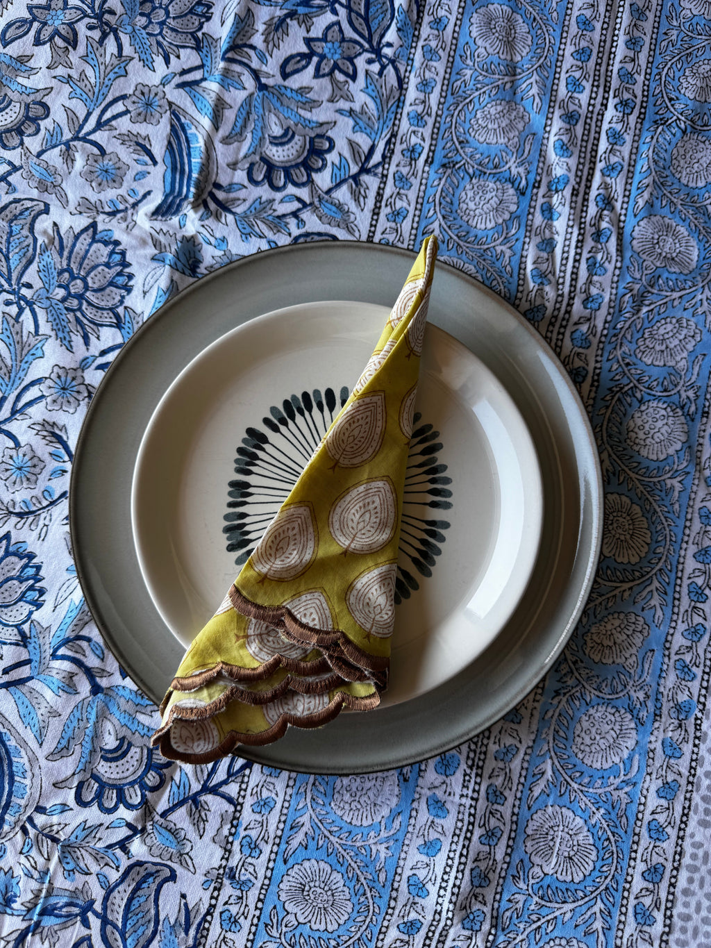A folded yellow fabric napkin with a beige and brown paisley pattern, placed on a white plate with a black circular motif, set on a gray plate over a blue and white patterned textile.