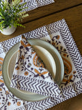 A styled table setting featuring a white linen napkin with a brown and purple floral paisley pattern and a gray and white chevron border, folded over a clear glass plate on a wooden table next to a small potted plant.