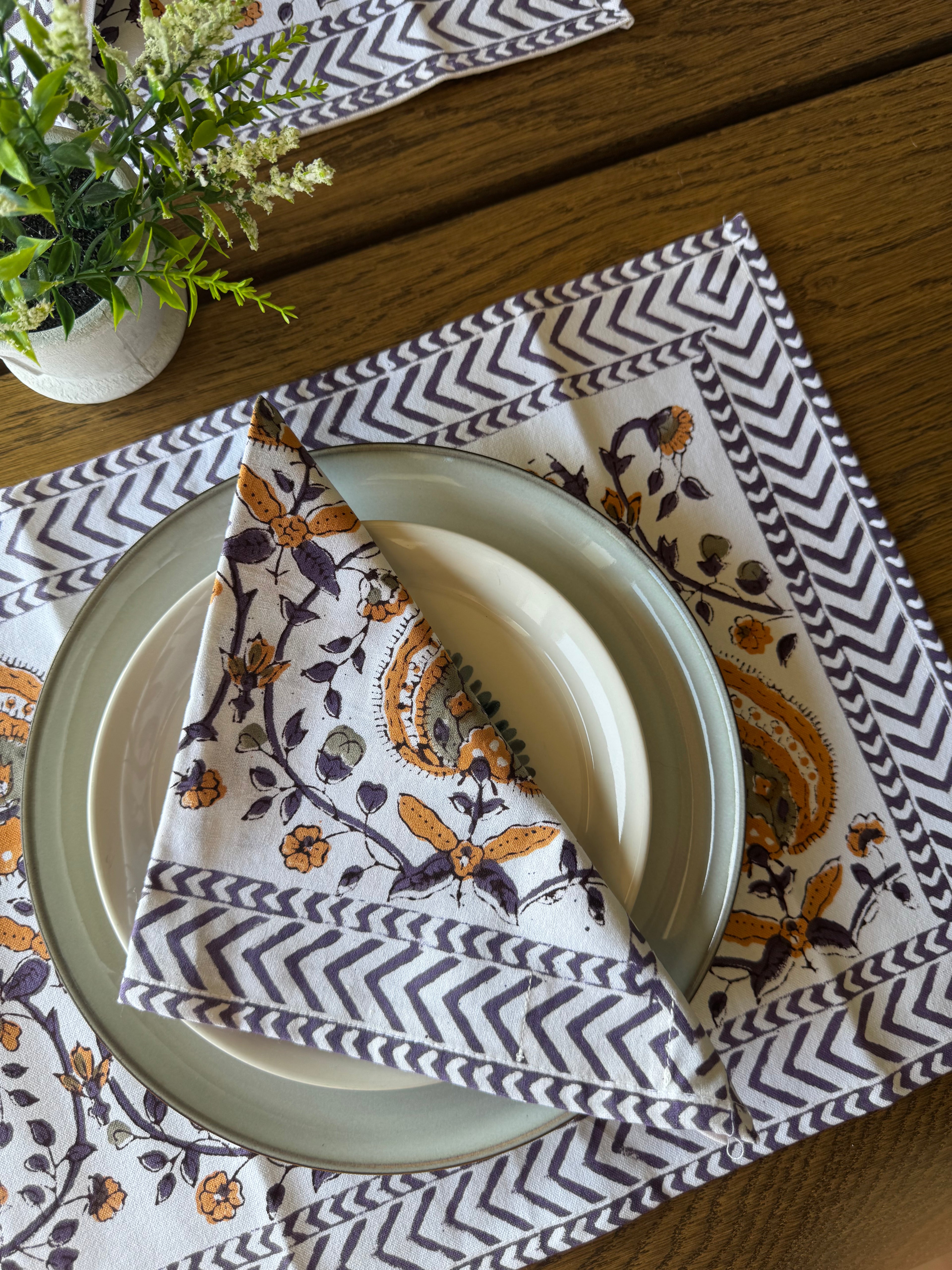 A styled table setting featuring a white linen napkin with a brown and purple floral paisley pattern and a gray and white chevron border, folded over a clear glass plate on a wooden table next to a small potted plant.