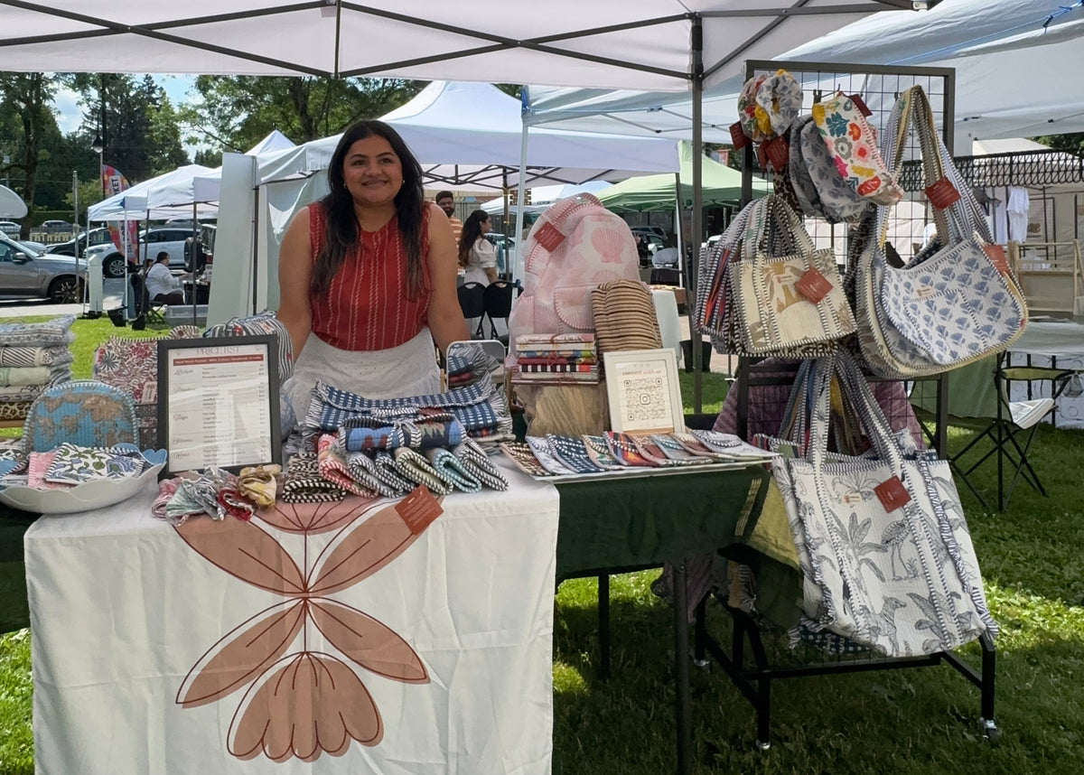 An outdoor market booth under a white canopy tent, with a woman standing behind a table displaying handmade goods. The table has a white cloth with the name 'Pehchān' and a large brown floral design, and nearby racks display patterned tote bags and other items.